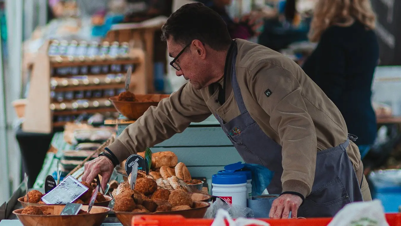 A baker in glasses and an apron arranges loaves on a market stall.